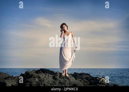 Pleine longueur de jeune femme barefootée tranquille en robe longue blanche touchant de longs cheveux sombres tout en se tenant sur la falaise rocheuse avec les yeux fermés près des vagues s Banque D'Images