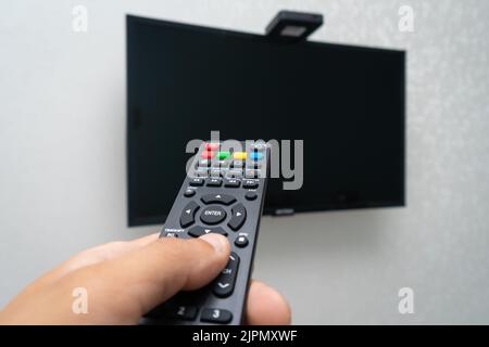 Gros plan d'une télécommande dans la main d'un homme dirigeant un téléviseur LCD noir suspendu sur un mur blanc Banque D'Images