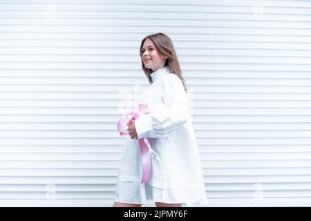 Joyeuse foire aux cheveux adolescente en robe blanche tenir rose boîte cadeau de vacances avec ruban. Près du mur blanc rayé Banque D'Images