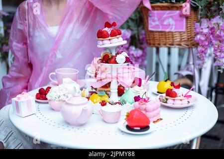 Une femme méconnaissable en imperméable rose mange des desserts aux baies sucrées, des gâteaux d'anniversaire, des bonbons aux fruits et du thé au café Banque D'Images