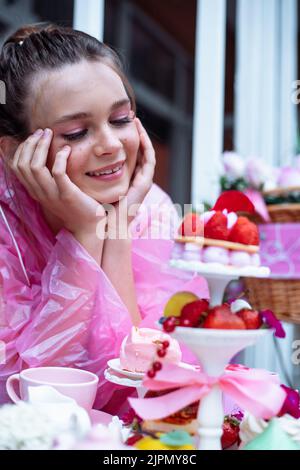 Portrait vertical en gros plan d'une adolescente rêvant en imperméable rose qui regarde les desserts sucrés sur la table Banque D'Images