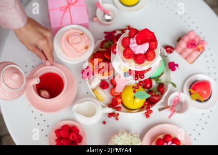 Une femme toute coupée verse du thé à la main dans une tasse près de la baie rose, des desserts aux fruits sur un support à gâteau sur une table blanche dans un magasin de bonbons. Haut Banque D'Images