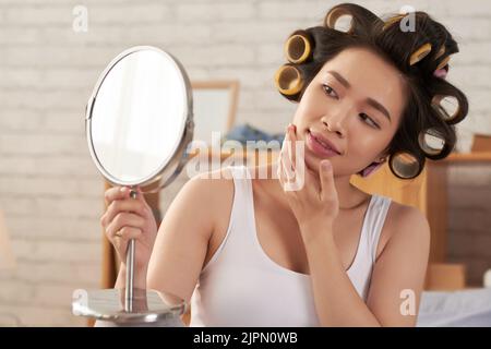 Jolie jeune femme asiatique avec des bigoudis regardant le miroir Banque D'Images