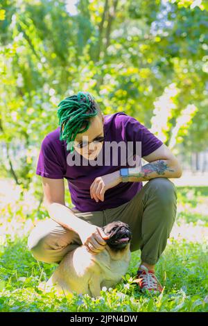 Un hipster avec des dreadlocks verts et des verres ronds noirs est assis sur une pelouse verte et de bourracher un pug mignon lors d'une journée ensoleillée d'été. Un homme marche un pug Banque D'Images