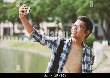 Homme asiatique gai portant des lunettes élégantes et portant un selfie sur un smartphone moderne, tout en profitant d'une agréable promenade dans un parc public, prise de vue portrait Banque D'Images
