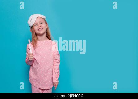 une petite fille en pyjama et un masque de sommeil montre une brosse à dents, sur un fond bleu Banque D'Images