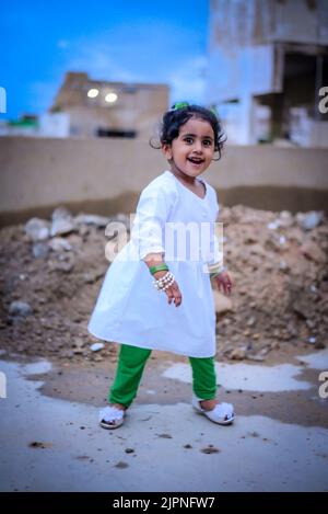 Un portrait d'une petite fille mignonne souriant et debout sur le sol en robe blanche et pantalon vert Banque D'Images