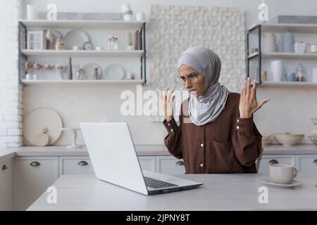 Belle femme musulmane à la maison dans hijab gris, travaillant à distance assis dans la cuisine avec un ordinateur portable, malheureux et déçu par le résultat Banque D'Images