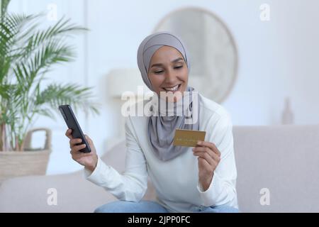 Belle femme musulmane assise sur un canapé à la maison en utilisant le téléphone et la carte de crédit bancaire pour les achats en ligne, femme dans hijab souriant et heureux Banque D'Images
