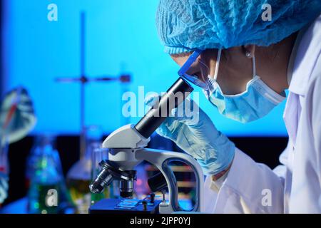 Vue en profil d'un microbiologiste asiatique concentré examinant l'échantillon à l'aide d'un microscope moderne tout en travaillant dans un laboratoire DIM Banque D'Images