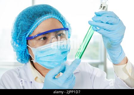 Joli chercheur asiatique portant un manteau blanc et un masque médical étudiant le contenu du tube à essai tout en travaillant dans un laboratoire moderne, portrait tourné Banque D'Images