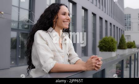 Caucasien hispanique 30s femme avec cheveux longs et bouclés femme d'affaires se tient sur la terrasse de balcon dans le bureau à la maison dans l'hôtel appréciant la contemplation Banque D'Images