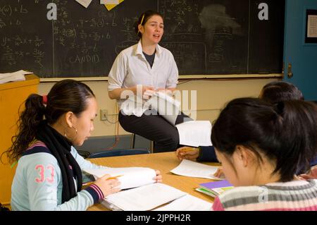 Éducation École secondaire enseignante parlant à la classe, crayon derrière son oreille, les étudiantes suivent le sujet avec des documents sur la table Banque D'Images