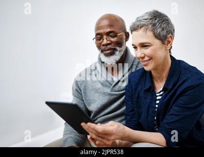 Es me montre toujours de nouvelles choses sur Internet. Un couple affectueux et expérimenté utilisant une tablette tout en se relaxant sur le canapé à la maison. Banque D'Images