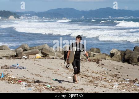 Un homme japonais plus âgé marche sur une plage parsemée de litière portant une planche de surf. Enoshima, Kanagawa, Japon. Banque D'Images