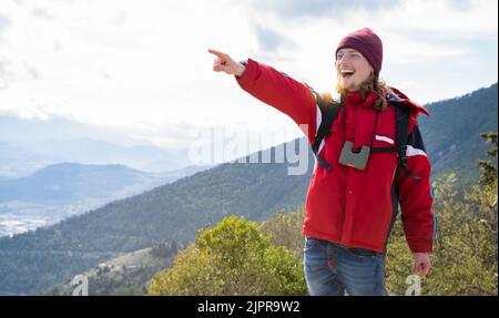 jeune randonneur vêtu de rouge avec un sac à dos sur le dessus de la montagne pointant de la main à l'horizon et souriant Banque D'Images