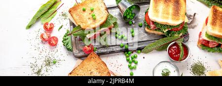 Sandwich au fromage, au jambon, à la tomate et à la salade fraîche, fait de pain de blé entier avec une croûte croustillante sur une planche de bois avec sauces sur fond blanc, Banque D'Images