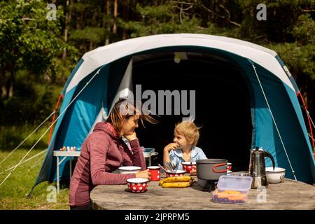 Famille, mère et enfant, prenant le petit déjeuner devant une tente dressée dans la forêt, tandis que camping sauvage en Norvège, en été Banque D'Images