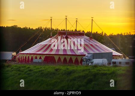 La tente de cirque à rayures au sommet de la colline verdoyante avant les bois au coucher du soleil Banque D'Images