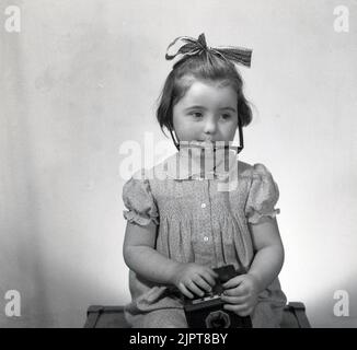 1963, historique, dans un studio photographique, une jeune fille, posant pour une photo amusante, dans les verres et le collier de sa mère et avec un joli ruban dans ses cheveux, tenant un appareil photo Conway film de l'époque, Stockport, Angleterre, Royaume-Uni. Banque D'Images