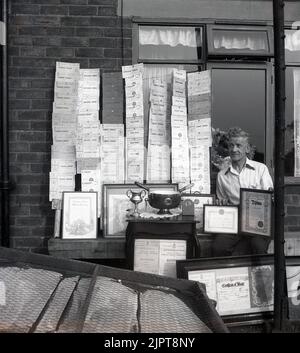 1955, historique, lauréat du prix, à l'extérieur de sa maison d'entreprise, un gentleman assis par le grand nombre de certificats et de trophées qu'il a gagné pour les compétences de jardinage, Stockport, Greater Manchester, Angleterre, Royaume-Uni. La plupart d'entre eux sont des prix pour le premier prix des spectacles de légumes, fruits et fleurs de la Corporation of Stockport. Banque D'Images