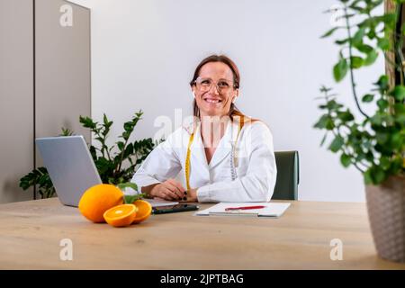 Portrait de jeune femme nutritionniste souriante dans son bureau en robe médicale avec des oranges fraîches. Dietologue faisant un plan de régime Banque D'Images