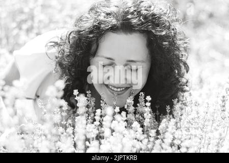 Portrait en gros plan noir et blanc de la jeune fille aux cheveux bouclés parmi les fleurs. Elle a fermé les yeux et apprécie l'arôme de la lavande. Sourire sincère. Détox numérique, plaisir de la vie lente. Mise au point sélective douce Banque D'Images