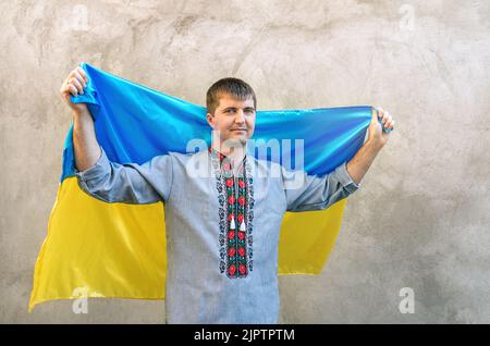 Le jeune homme porte le drapeau bleu-jaune de l'Ukraine au-dessus de sa tête. Homme dans une chemise brodée traditionnelle sur fond de mur en béton texturé. Vue de face, espace de copie. Jour de l'indépendance de l'Ukraine Banque D'Images