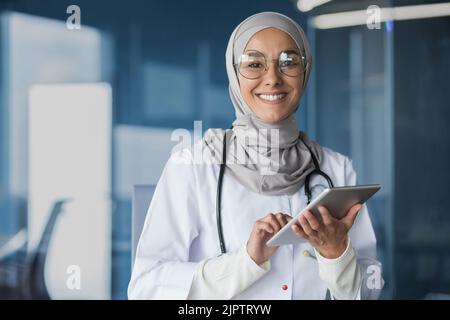 Jeune belle femme asiatique musulman médecin dans hijab souriant et regardant la caméra travailler avec un ordinateur tablette dans le bureau de clinique moderne à l'intérieur du médecin dans une robe médicale et des lunettes. Banque D'Images