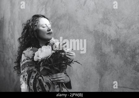 Photo noire et blanche d'une fille aux yeux fermés tenant un bouquet de pivoines. Femme dans une robe brodée, de longs cheveux bouclés rouges sur fond de mur de béton. Jour de l'indépendance Ukraine. Mise au point douce Banque D'Images