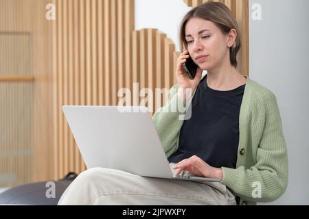 Femme d'affaires sérieuse discutant des détails du projet via un téléphone portable tout en travaillant à distance sur un ordinateur portable Banque D'Images