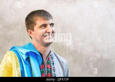 Portrait rapproché du jeune homme avec drapeau bleu et jaune de l'Ukraine. Homme dans une chemise brodée traditionnelle sur fond de mur gris en ville. Vue de face, espace de copie. Jour de l'indépendance de l'Ukraine Banque D'Images