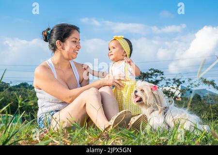 latina mère assise sur l'herbe avec son beau bébé brunette et son chien Shih tzu, dans un terrain plat entouré d'arbres. Fond de bleu summ Banque D'Images