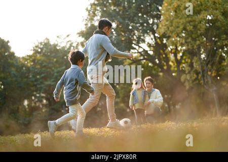 père et fils asiatiques jouant au football à l'extérieur dans le parc tandis que la mère et la fille regardent sur le côté Banque D'Images