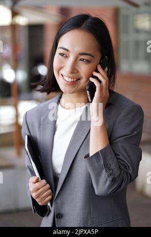 Jeune femme d'affaires asiatique souriante parlant sur un téléphone portable debout à l'extérieur. Banque D'Images