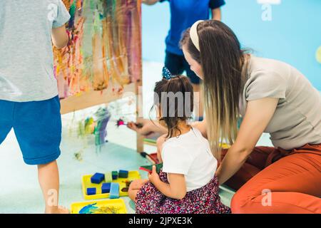 Peinture de film adhésif. Peinture pour tout-petit avec une éponge, des pinceaux et des peintures sur un film adhésif enroulé autour de l'étagère en bois. Un enseignant les aidant. Activité créative pour les enfants habiletés sensorielles et développement visuel perceptif à la maternelle. Photo de haute qualité Banque D'Images