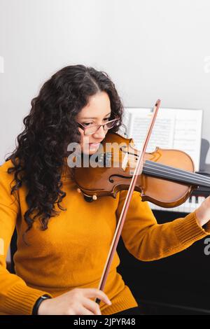 Femme brune souriante portant un chandail jaune, et jouant du violon en lisant de la musique en feuilles. Banque D'Images