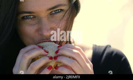 L'été, à l'extérieur. Portrait de la belle femme brune, avec des cheveux foncés, avec de l'Apentite manger fastfood, le hamburger à morsure sexuelle. Elle a des lèvres Chubby. Photo de haute qualité Banque D'Images