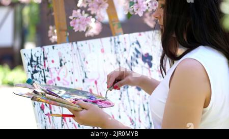 une belle femme peintre en robe blanche, en fleurs de verger de pomme de printemps, elle mélange les peintures avec une petite spatule spéciale sur la palette. l'artiste peint une image de fleurs. Photo de haute qualité Banque D'Images