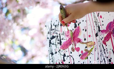 gros plan, femme main, peintre, artiste peint une image de fleurs dans le verger de pomme de printemps en fleur, elle applique des peintures sur toile avec une petite spatule spéciale, tient la palette avec des peintures. Photo de haute qualité Banque D'Images