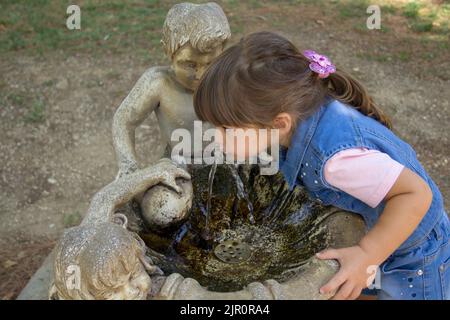 Image d'une adorable petite fille qui boit de l'eau provenant d'une fontaine décorée dans un parc public. Jeux et vie en plein air avec les enfants pendant l'holida Banque D'Images