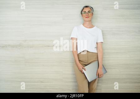 Femme d'affaires mûre souriant gaiement tout en se tenant contre un mur dans un bureau. Femme entrepreneur prospère tenant un ordinateur portable dans un poste de travail moderne Banque D'Images