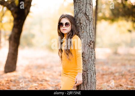 Mignon élégant enfant fille 4-5 ans vêtements tricoté jaune robe et lunettes de soleil posant dans le parc extérieur. Saison d'automne. Enfance. Banque D'Images