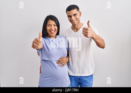 Un jeune couple hispanique attend un bébé qui se tient debout sur l'arrière-plan pour approuver le geste positif avec la main, les pouces vers le haut souriant et heureux pour le succès. w Banque D'Images