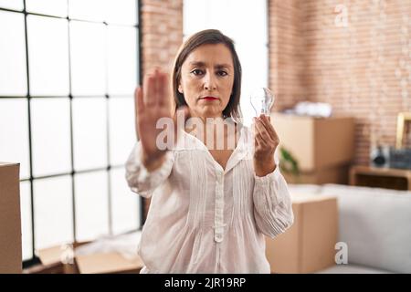 Femme hispanique d'âge moyen tenant une ampoule pour l'inspiration et l'idée avec la main ouverte faisant stop signe avec une expression sérieuse et confiante, la défense gges Banque D'Images