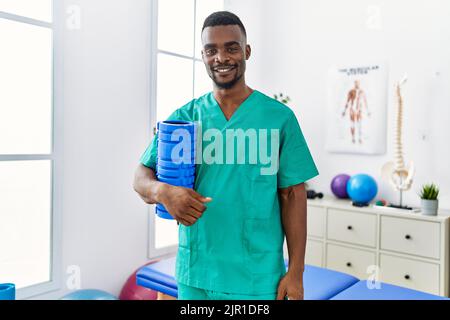 Un jeune physiothérapeute africain tenant un rouleau en mousse à la clinique a l'air positif et heureux debout et souriant avec un sourire confiant montrant le tee Banque D'Images