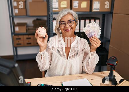 Femme d'âge moyen avec des cheveux gris travaillant à la petite entreprise ecommerce tenant la banque de piggy et zloty souriant avec un sourire heureux et frais sur le visage. Montrant Banque D'Images