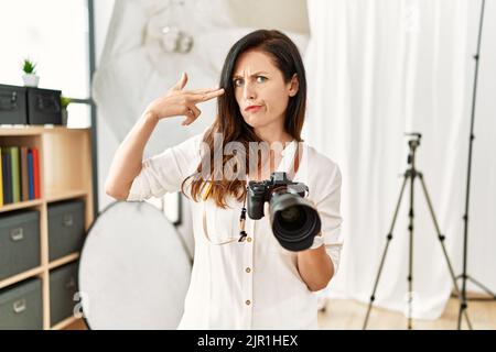 Belle femme caucasienne travaillant comme photographe au studio de photographie de tournage et de se tuer pointant la main et les doigts à la tête comme un fusil, suici Banque D'Images