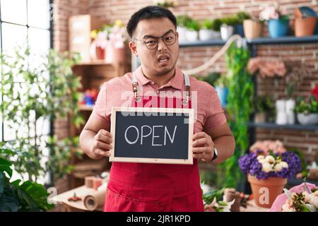 Jeune homme chinois travaillant chez un fleuriste avec un signe ouvert, expression sans indice et confuse. Notion de doute. Banque D'Images