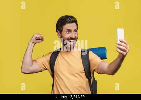 Homme excité avec des équipements touristiques prenant selfie sur smartphone sur fond jaune studio, faisant la photo de lui-même Banque D'Images